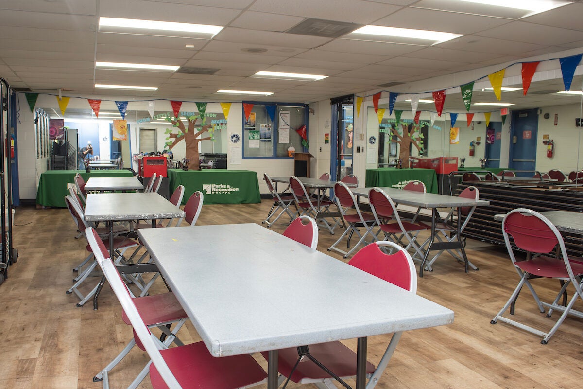 A multipurpose room with mirrors on several of the walls, containing six folding tables with four chairs each. At the far wall are two long tables with green Parks and Rec tablecloths, as well as a large, paper decoration of a tree. Decorative flag garlands hang along the tops of the walls.
