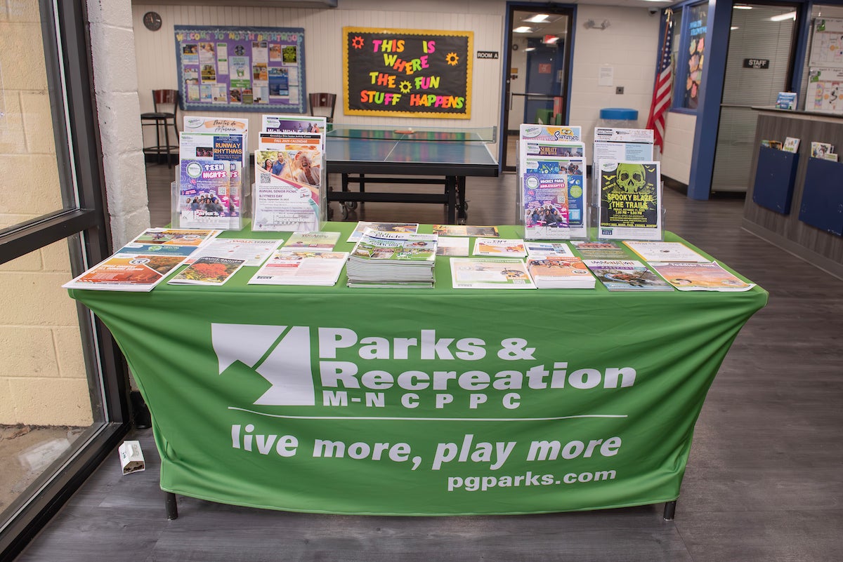 Inside a lobby area, a table with a green Parks and Rec tablecloth contains dozens of neat stacks of flyers, program guides, and literature. In the room behind this is a ping-pong table and a bulletin board; to the right, the edge of a reception desk is just visible.