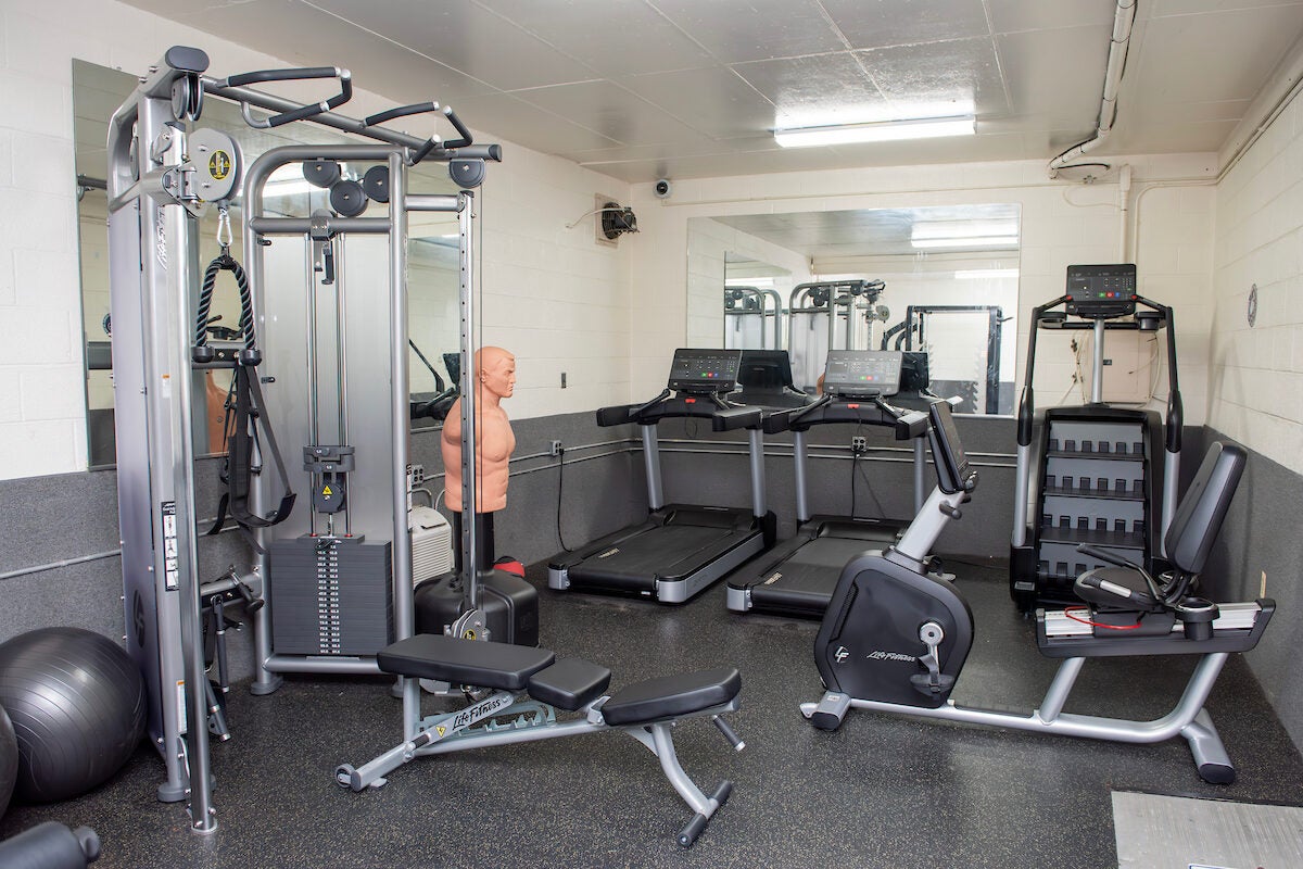 A fitness room with white and gray walls, mirrors on the walls, and multiple pieces of exercise equipment, including treadmills, a stair-climber, a boxing dummy, and a weight machine.