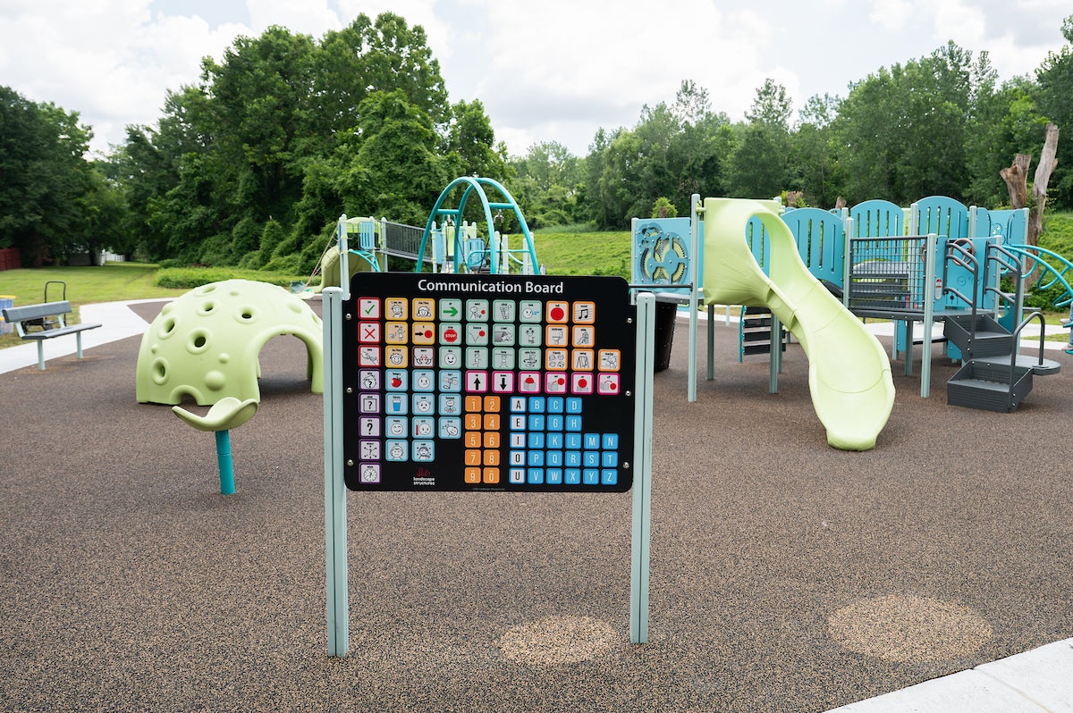 At an outdoor playground, a Communication Board is installed near the edge, containing various icons and images as well as the alphabet and numbers. The playground features slides, platforms, monkey bars, spinning elements, swings, rope webs, and more.