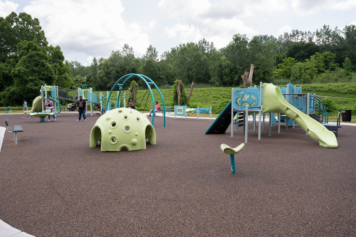 An outdoor playground featuring spinning elements, slides, swings, platforms, rope webs, climbing walls, musical instruments, and more.
