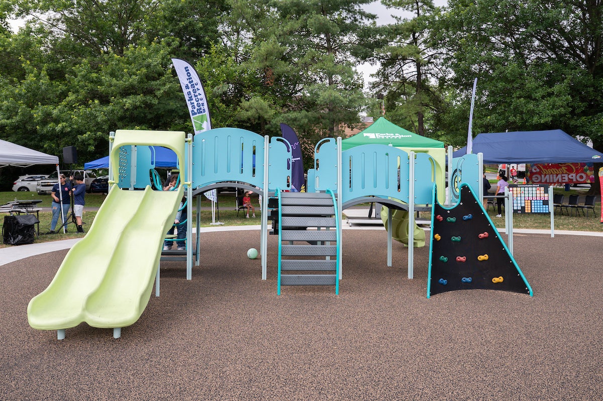 A play structure at an outdoor playground, featuring slides, a climbing wall, bridges, platforms, monkey bars, and more. Beyond the edge of the playground, event tents are set up in the grass under some tall trees.