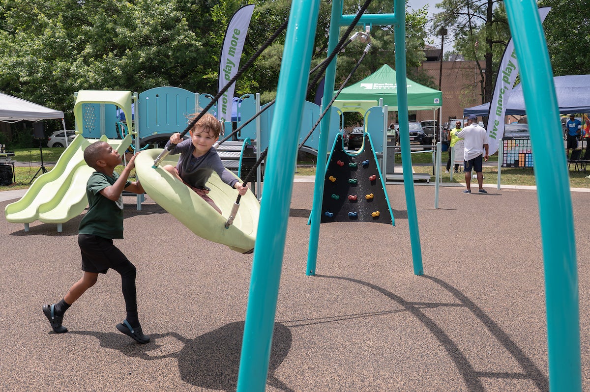 At an outdoor playground, one boy pushes another on a large, plastic, dish-style swing. Behind them is a large play structure with slides, platforms, and a climbing wall. Beyond the playground, several event tents are set up in the grass.