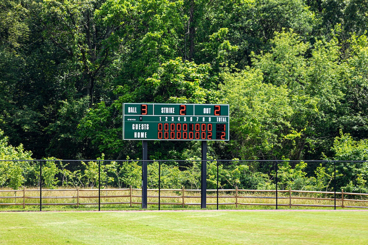 A scoreboard installed at the fenced outfield edge of a baseball diamond. Beyond the fence are tall tees.