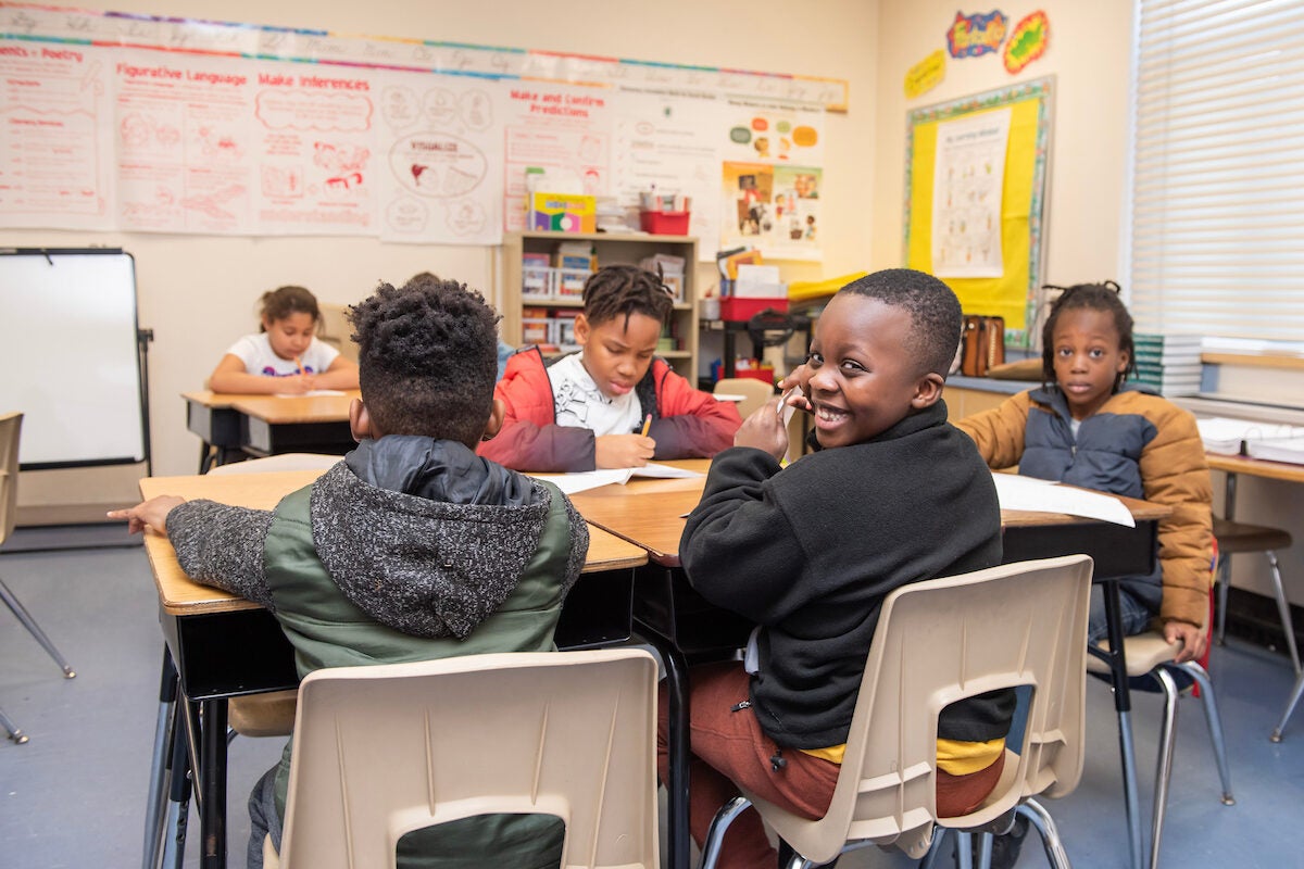 In a classroom, four boys sit at school desks writing with pencils in workbooks. One boy whose back is to the camera has turned around and is giving the photographer a mischievous grin.