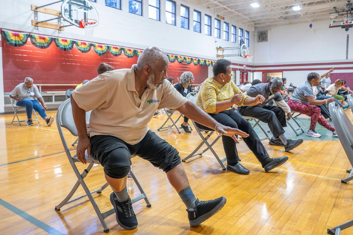 In a gymnasium, seniors sit in rows of folding chairs doing seated strength exercises. They are all extending their left legs and arms while looking at the class instructor.