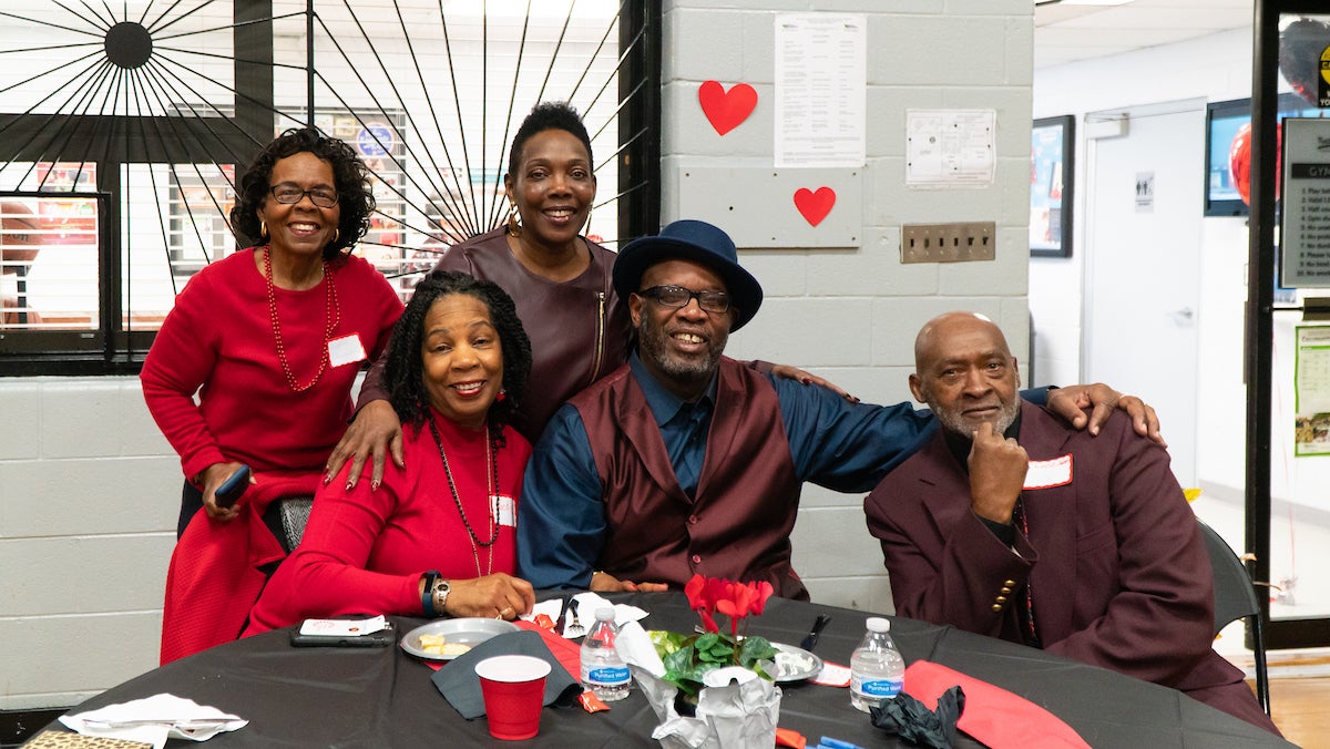 In a gymnasium decorated for Valentine's Day, five smiling adults in fancy outfits pose at a table with red flowers in the middle. They are all wearing red or burgundy outfits for the Valentine's theme.
