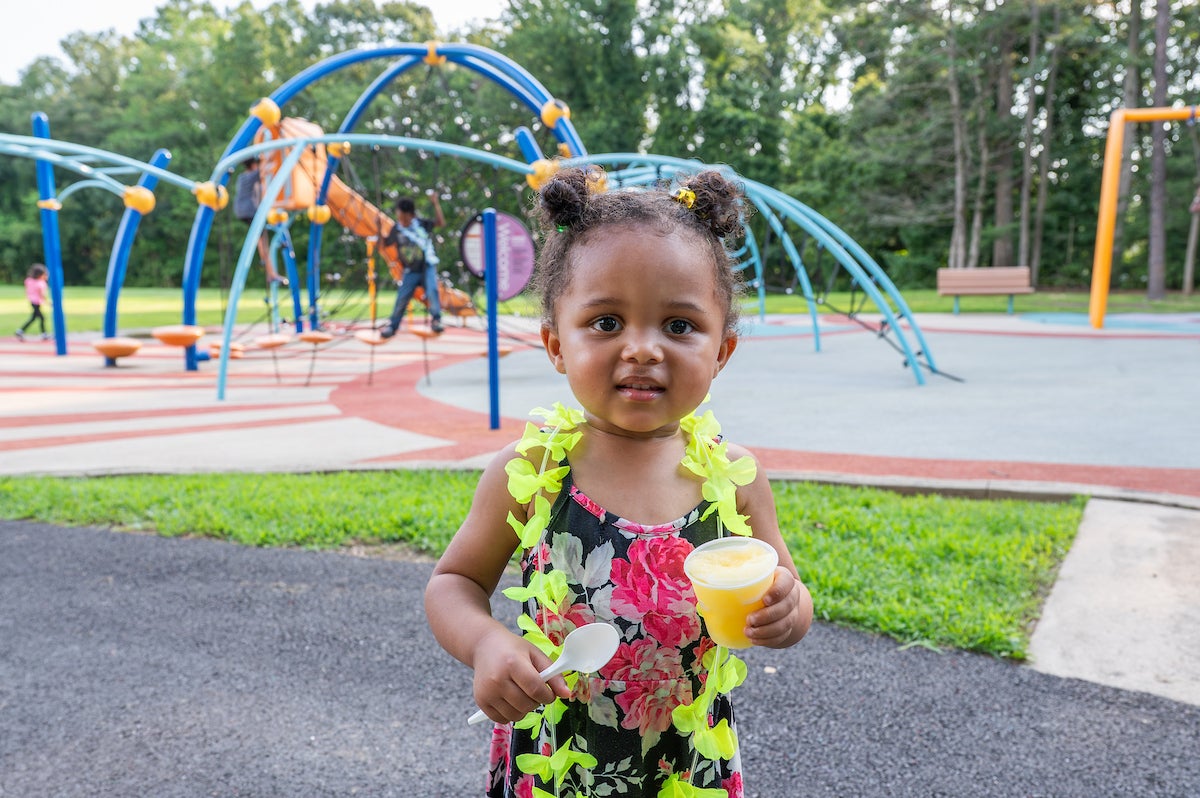 A toddler holds a frozen treat while wearing a yellow lei-style flower necklace. Behind her is a large, colorful, outdoor playground.