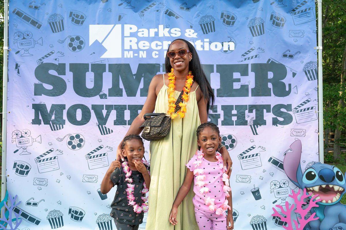 A mom poses with her two small children in front of a banner reading PARKS AND RECREATION SUMMER MOVIE NIGHTS. They all wear colorful flower lei-style necklaces. A cut-out character from the movie Lilo and Stitch is positioned in the right corner.