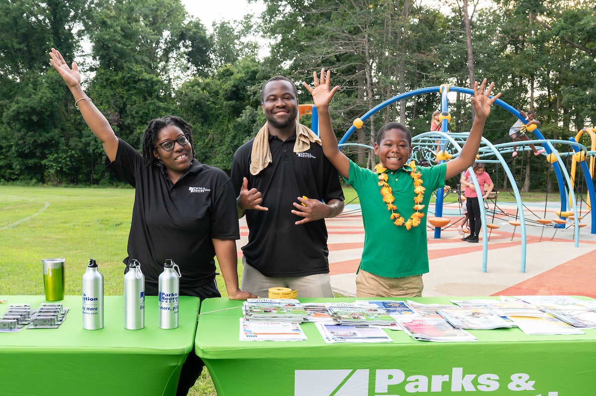 Two adults in black Parks and Rec staff shirts, and a boy in a green shirt, smile and wave while stationed behind two long information tables with green Parks and Rec tablecloths. The tables contain stacks of flyers, program guides, and giveaways. Behind them is a large, colorful outdoor playground.