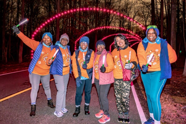 Six smiling women wearing matching orange holiday sweatshirts along with vests, hats, and gloves, pose on a paved road at night. Behind them are multiple arches over the road made of holiday lights. It is night, and the woman all hold glowing sticks.