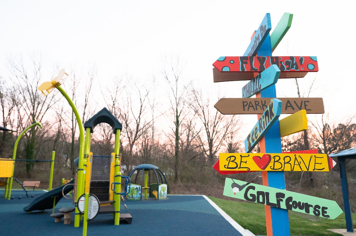 Near a green and blue outdoor playground is a wooden signpost with eleven hand-painted directional signs nailed around it at different heights. The arrow signs include locations like GOLF COURSE, ICE RINK, and PARK AVE along with motivational phrases like FLY HIGH and BE BRAVE.