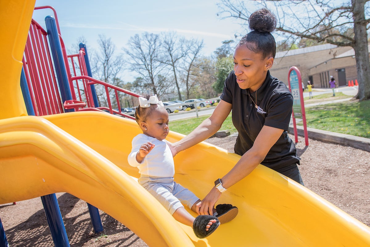 A woman wearing a black staff shirt holds onto a small toddler, helping her to go down a yellow plastic slide on an outdoor playground. Kids wearing bunny-ear headbands play in the grass nearby.