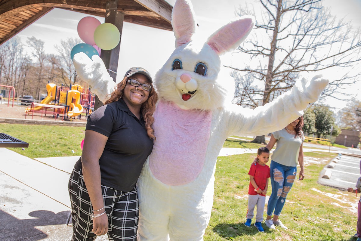 A smiling woman poses with her arm around a costumed Easter bunny. They are standing next to a picnic shelter decorated with pastel balloons; behind them is a colorful playground.