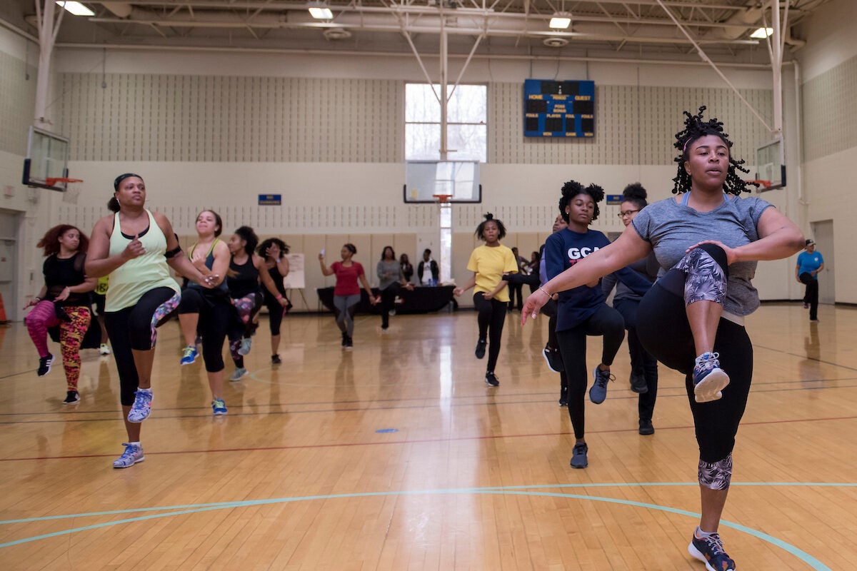In a gymnasium, several dozen women and teens in athletic clothing exercise in a Zumba class.