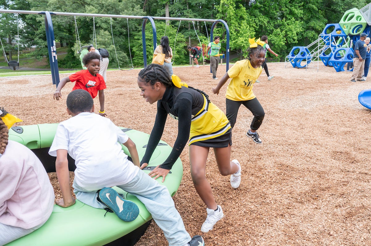Several children run around near a green spinning ring feature at an outdoor playground. Behind them are swings and a large, blue and green climbing structure.