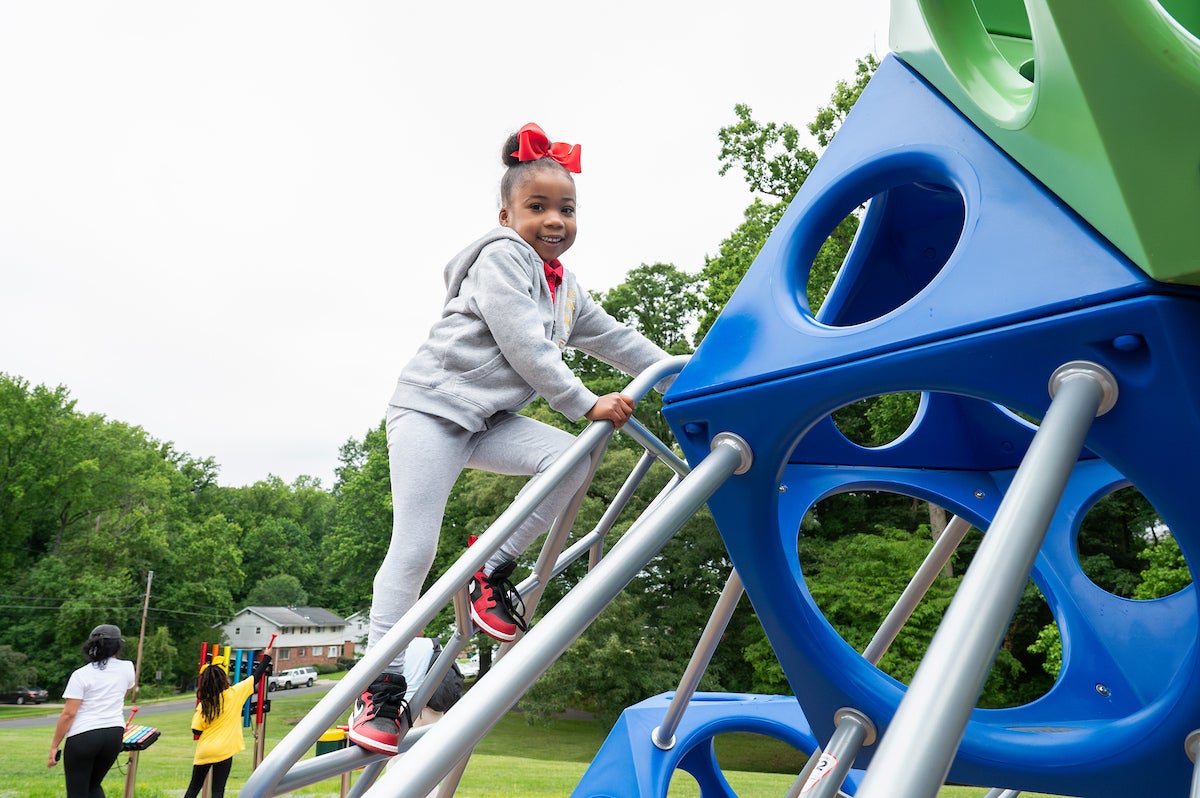 A child wearing gray sweats, a red shirt, a red bow in her hair, and red, black, and white Nike shoes climbs on a large play structure at an outdoor playground. Behind her, a girl is playing a colorful xylophone at the edge of the playground.