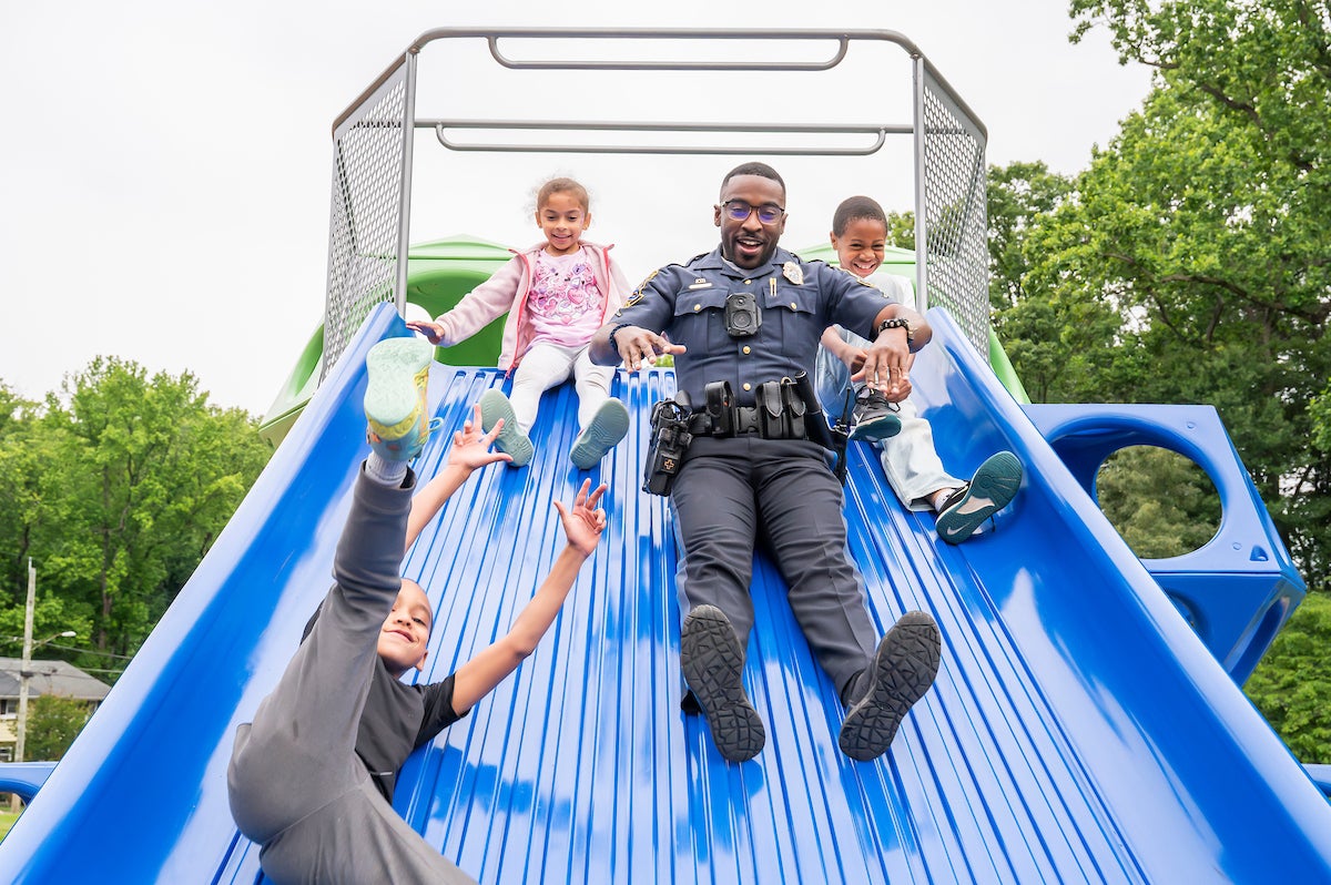 A male police officer and three children smile while sliding down an extra-wide, blue plastic slide at an outdoor playground.