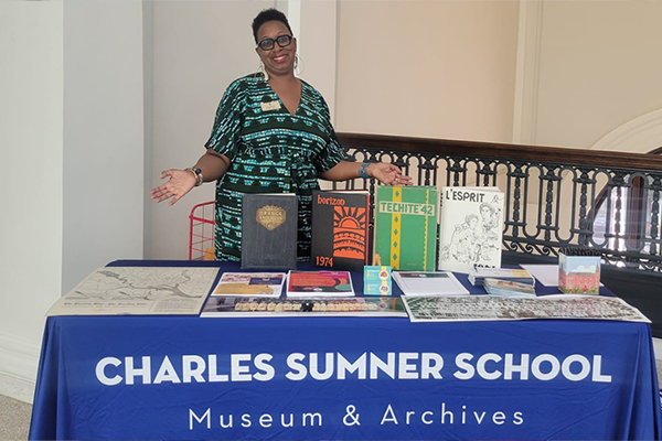 A smiling woman in a patterned black and teal dress stands behind an information table displaying books, maps, flyers, and literature. Behind the table is an ornate wooden bannister. A blue tablecloth covering the table says CHARLES SUMNER SCHOOL MUSEUM & ARCHIVES on the front.