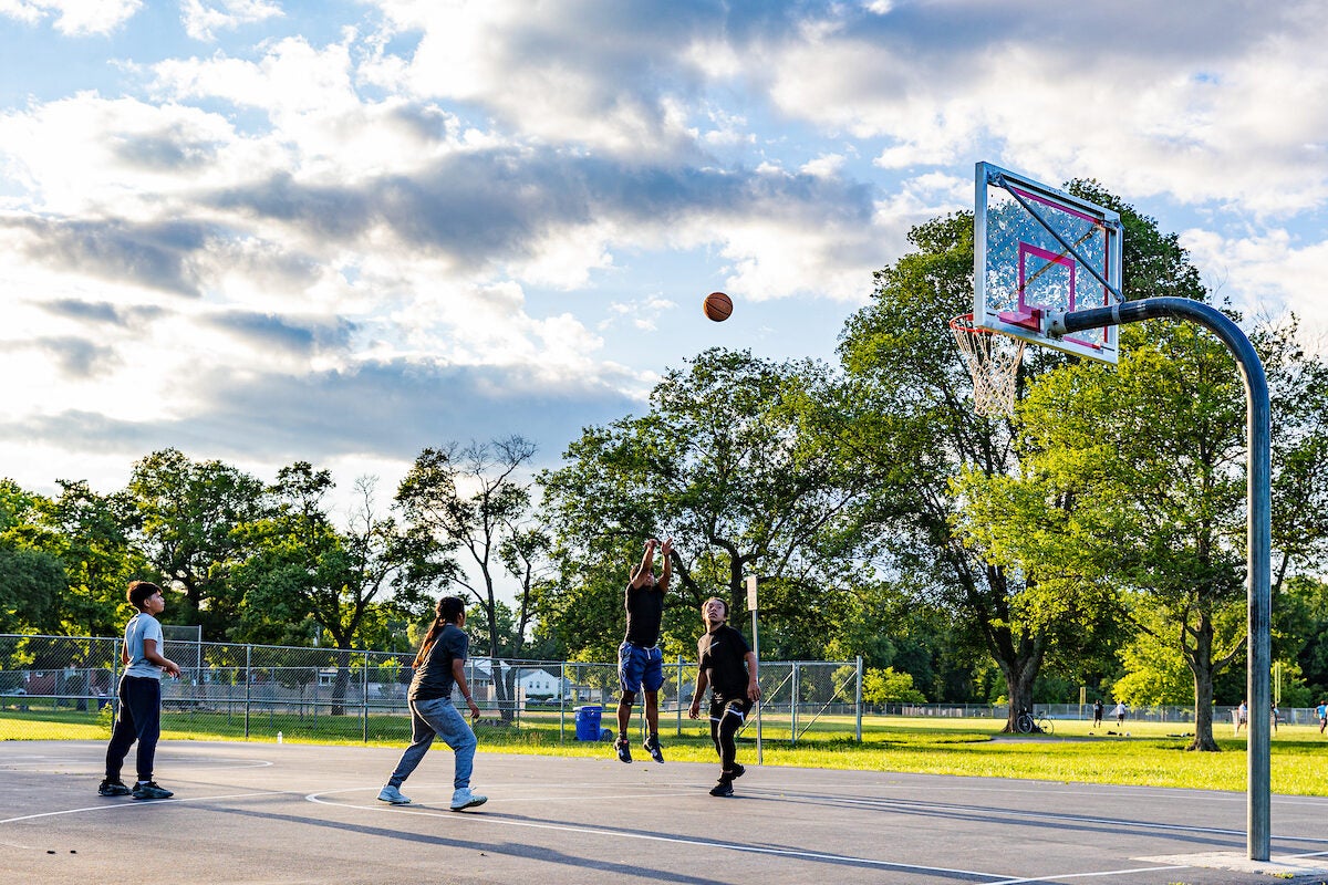 On a sunny day, four boys play basketball on an outdoor court. One boy is about a foot off the ground with his arms up, having just thrown the ball arcing toward the goal.