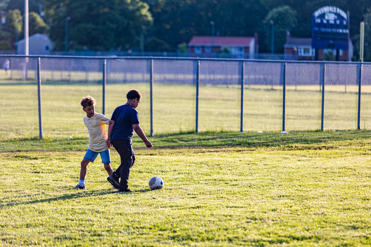 In a grassy field near a chain-link fence, two boys kick a soccerball. In the distance is a large scoreboard with the words RIVERDALE PARK across the top. Several single-family homes are visible in the distance.
