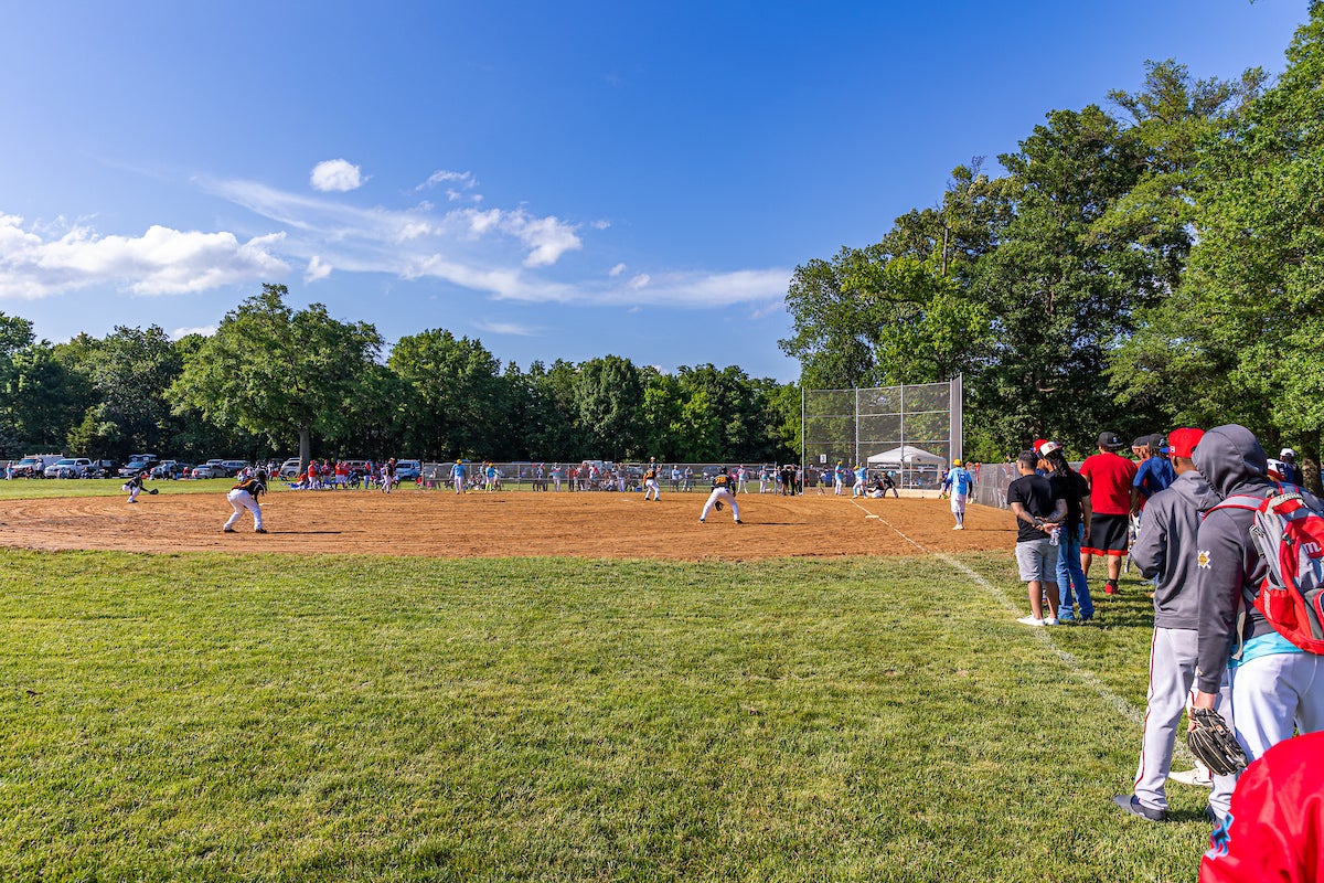 On a sunny day, a baseball game is being played on a baseball diamond in the sun. One team wears blue jerseys and the other wears black. There are trees in the distance. The photo is taken from the outfield near the third baseline.
