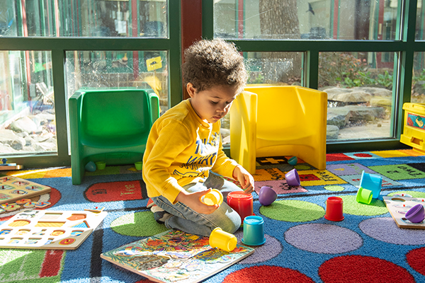 A small child plays with stacking cups on a colorful rug next to large windows. Near the child are wooden puzzled and plastic youth chairs.
