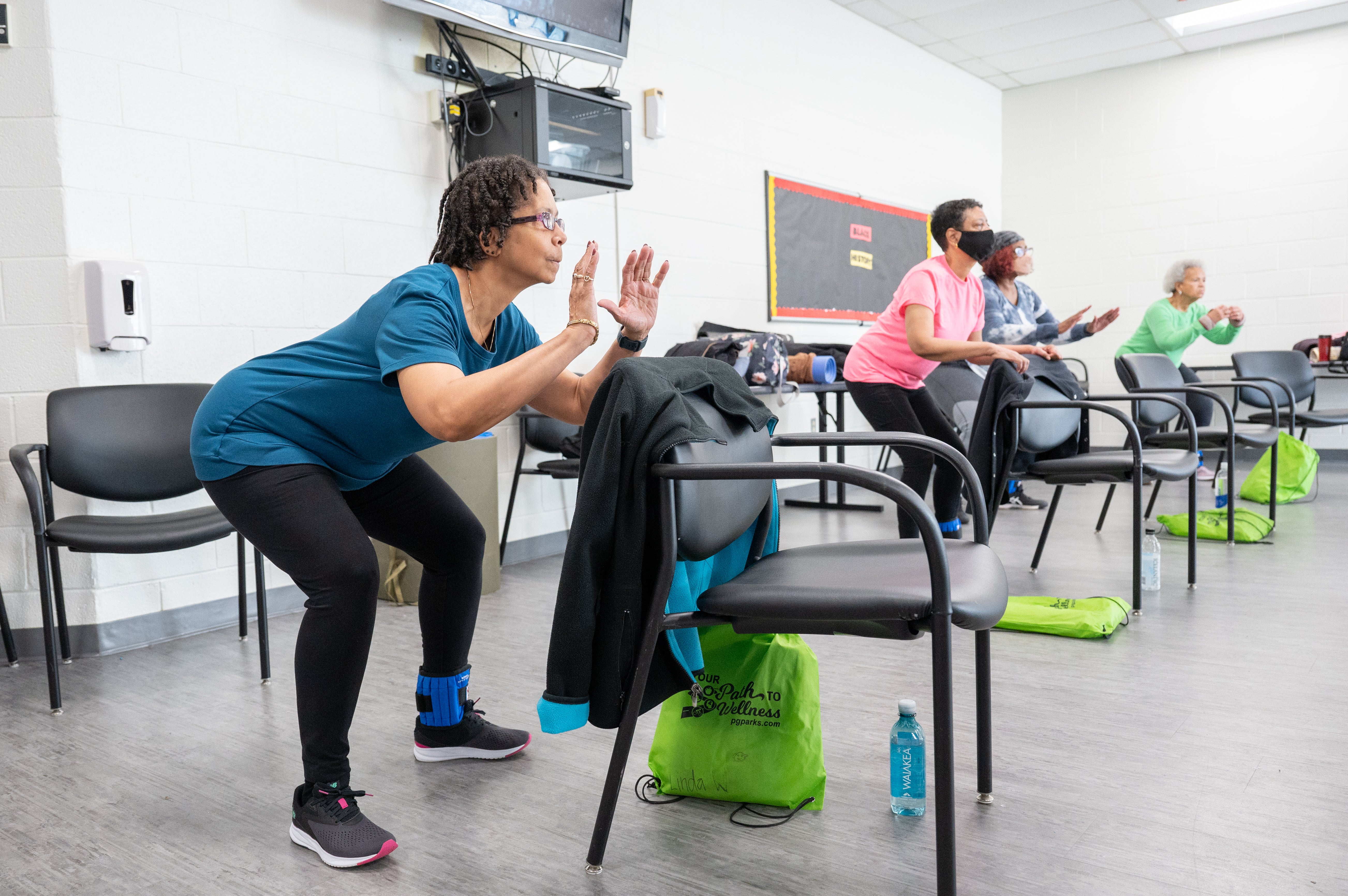 In a white room with gray flooring, four senior women wearing exercise clothes crouch behind black chairs with their arms slightly raised, following along with an unseen fitness instructor. There are green drawstring bags on the floor by each chair with the words "Our path to wellness" printed on them.