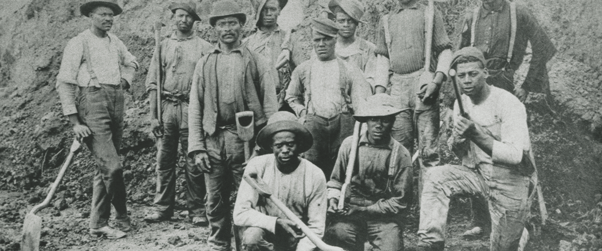 Black-and-white historical photo depicting about a dozen Black workmen posing with shovels and pickaxes. They appear to be in a deep hole or trench.
