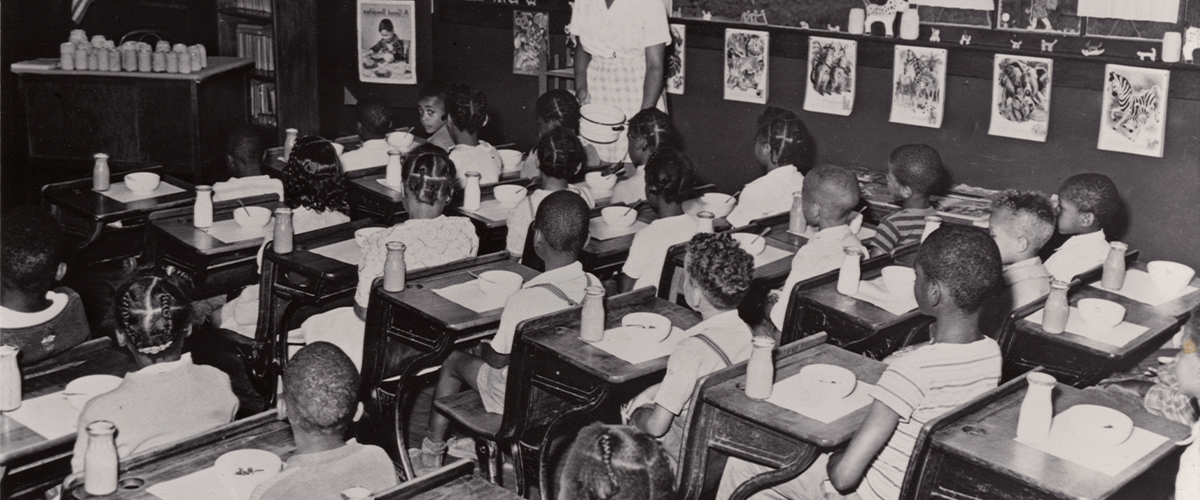 Black-and-white historical photo of a classroom of Black children sitting at old-fashioned desks. Each child has a bowl of food and a small bottle of milk on their desk. A teacher stands at the far wall.