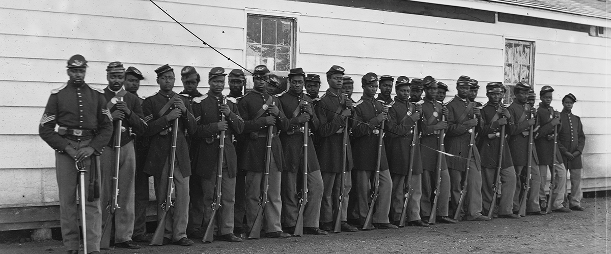 Black-and-white, historical photo depicting about 30 Black soldiers in uniforms, standing in two rows next to a white building. The men in the front row hold rifles.