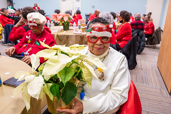 A senior woman wearing novelty holiday glasses holds a white poinsettia plant and smiles. She is seated at a table in a room filled with other seniors, also sitting at tables and wearing red holiday clothing and novelty holiday glasses.
