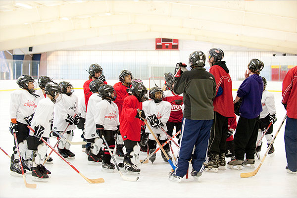 A group of boys wearing ice hockey uniforms gathers around three adults on the ice at an indoor ice rink. Most of the boys wear white hockey sweaters with the word Capitals on the front. They all wear black helmets and hold wooden hockey sticks.