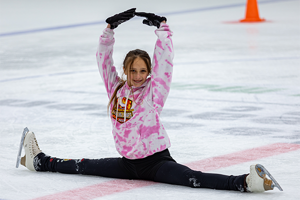 A girl wearing ice skates does the splits with her held arms in a dancer's arabesque pose, on the ice in an indoor ice rink. She wears a pink tie-dyed hoodie and black gloves.