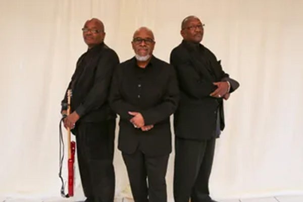 Three men wearing glasses and black suits pose together, standing shoulder-to-shoulder. One holds a red electric guitar.