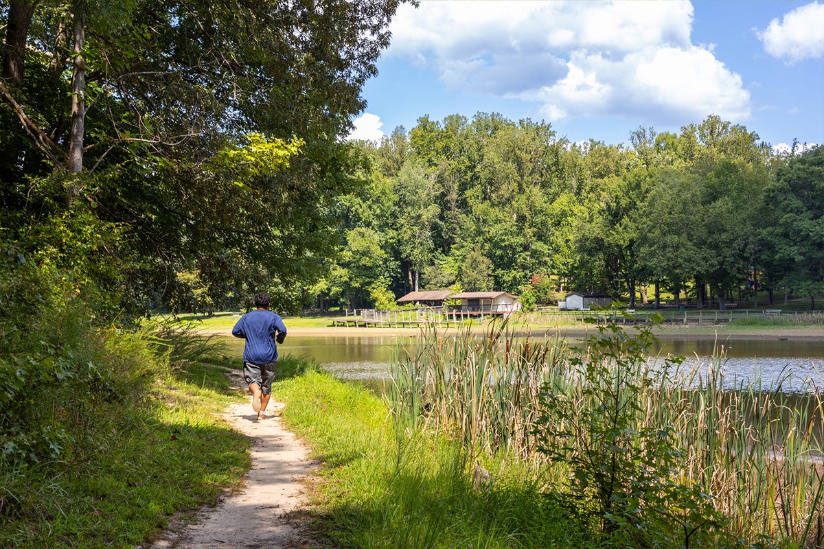 A person jogs on a natural surface trail that runs along the bank of a body of water.