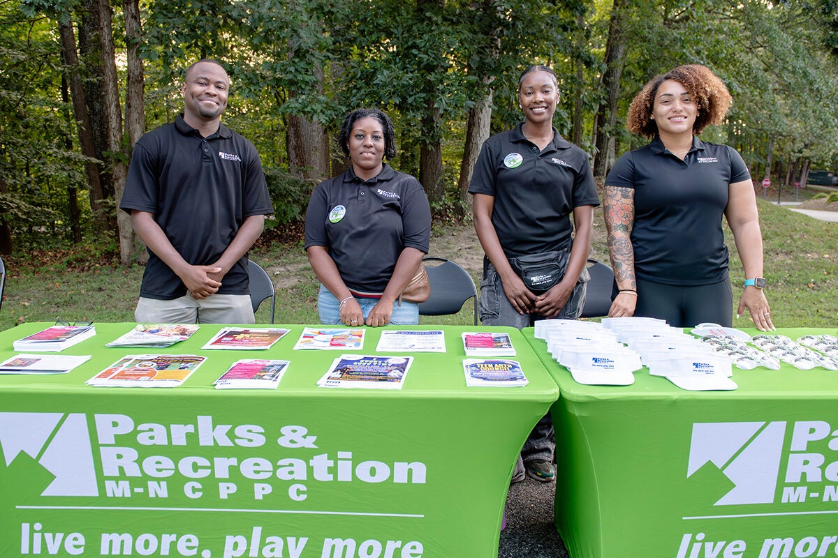 Four smiling staff members stand behind two information tables set up in the grass in a park. The tables contain flyers and giveaway items.