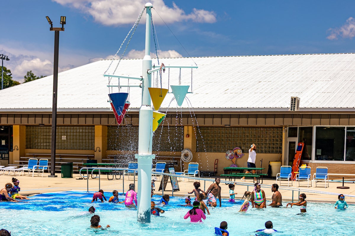 People enjoy a large outdoor pool with a central splash feature, next to a large, tan building.
