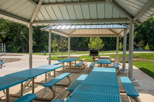 A row of blue tables under two metal picnic shelter rooves. There are plenty of trees, and a playground off to the side.