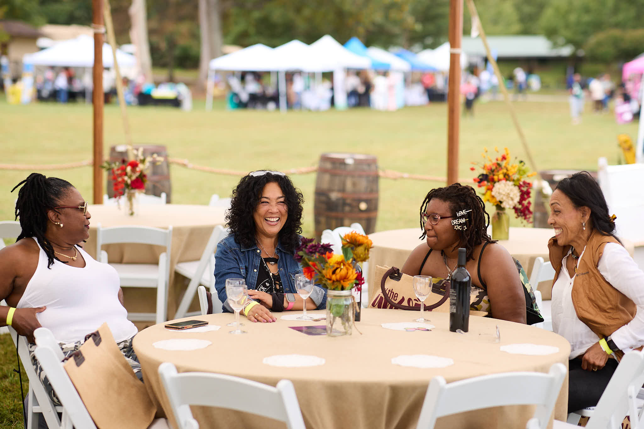 People seated around a decorated table at an outdoor community event, with tents, flowers, and open lawn in the background.