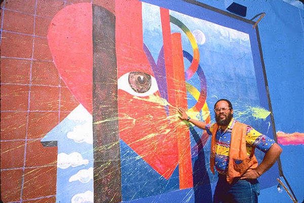 A man in a colorful shirt poses next to a large, colorful, abstract mural.