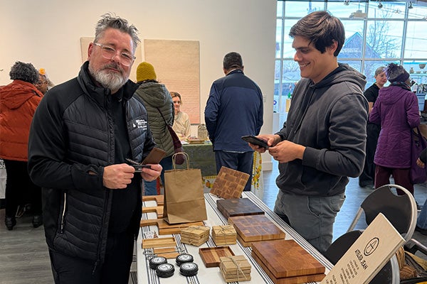 A happy customer buys something from a vendor at a craft fair, next to a table displaying artisan wood cutting boards and coasters.