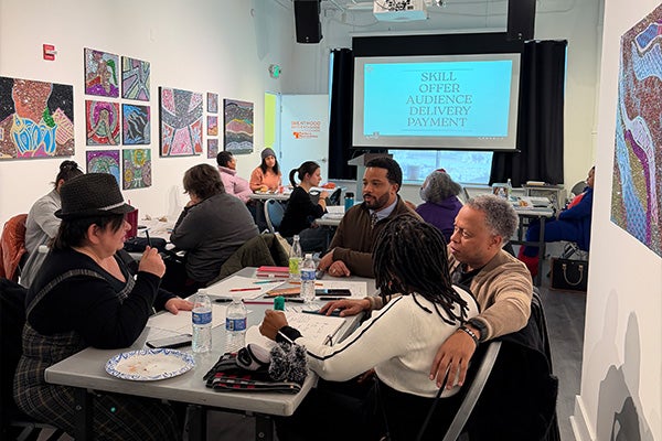 Adults sit around long tables in an art gallery, participating in a workshop. A slide projected on a screen lists the words Skill, Offer, Audience, Diversity, Payment.