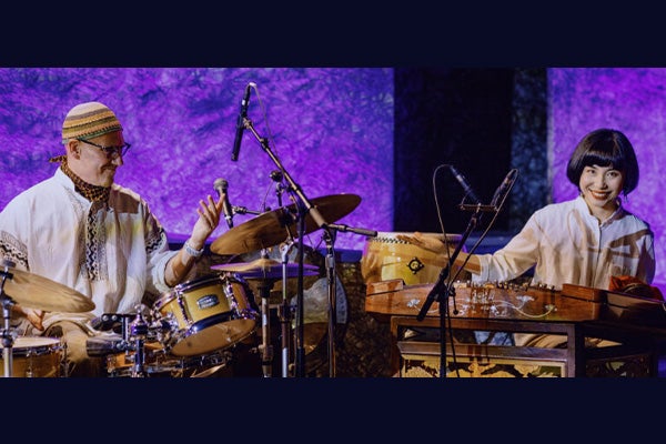 A musical performance by a man sitting behind a drum kit and a woman sitting behind a large Chinese dulcimer.
