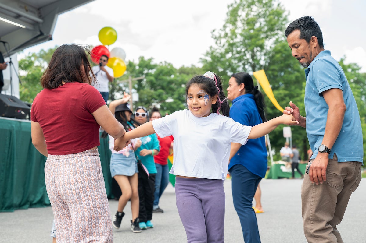 Child with face paint holds hands while dancing with adults at an outdoor community event near a stage.