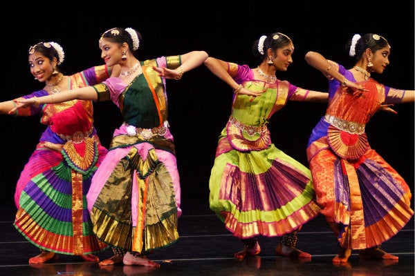 Four dancers perform on a stage wearing colorful Asian costumes with pleated skirts.
