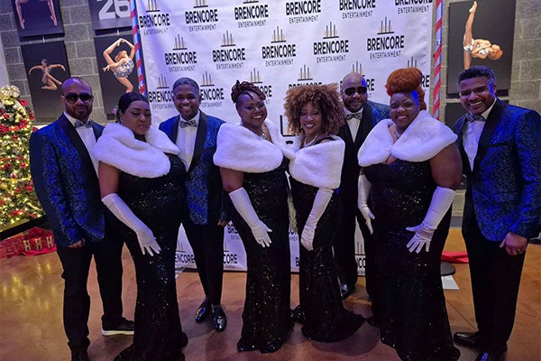 Eight men and women in formal wear pose smiling. The men wear blue brocade jackets and the women wear white fur wraps and long gloves.
