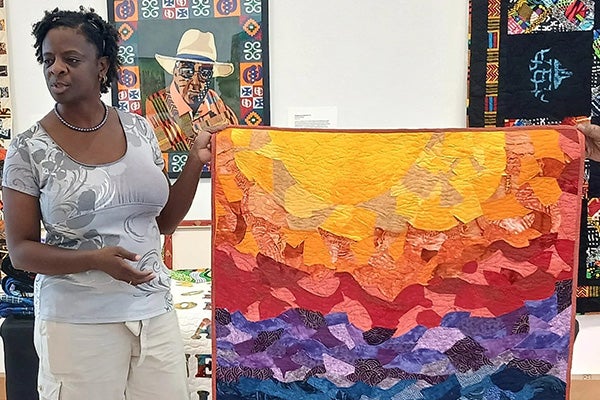 A woman holds up a colorful, quilted, abstract artwork in an art gallery.