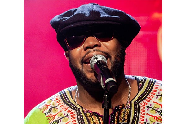 A man in a colorful tunic, black hat, and sunglasses sings into a microphone.
