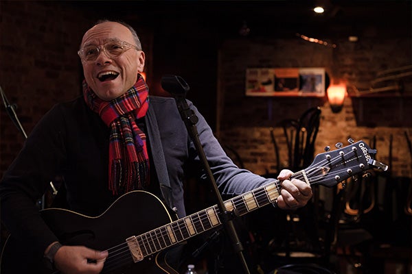 A man in a red plaid scarf singing and playing guitar.